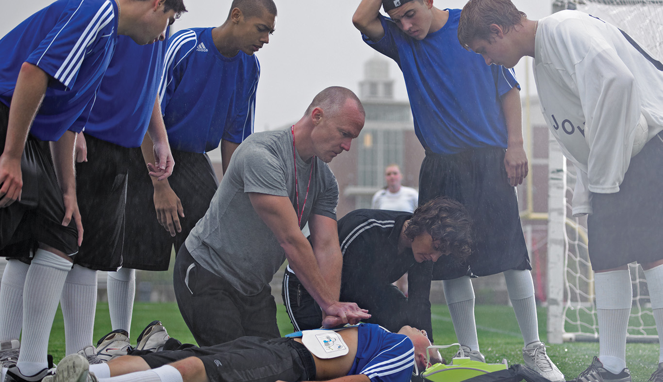 Coach demonstrating an AED on a student in front of teammates