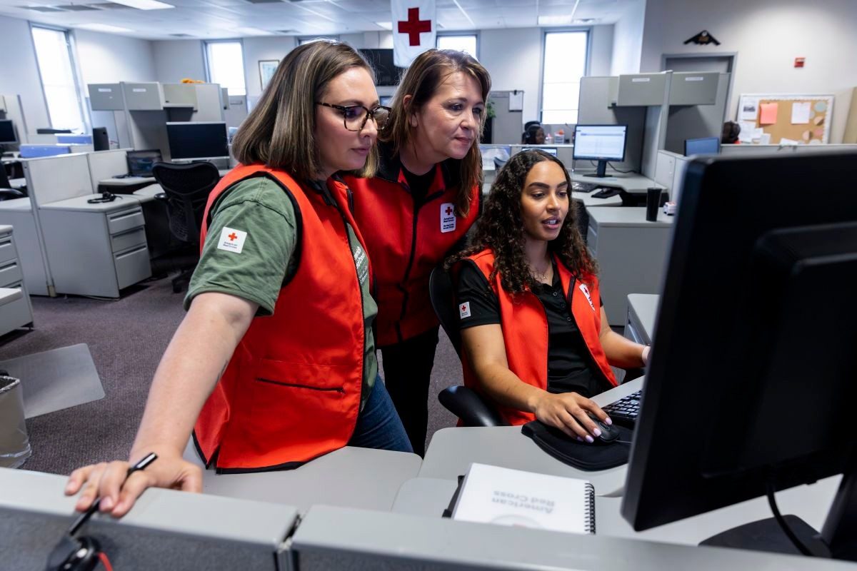 Three Red Cross volunteers sit and stand around a desk while working on a computer.