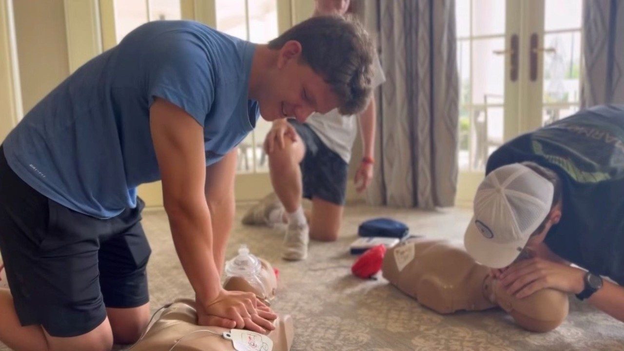 a young man in a blue tee shirt is leaning over a CPR dummy and practicing chest compressions. other learners are practicing nearby.